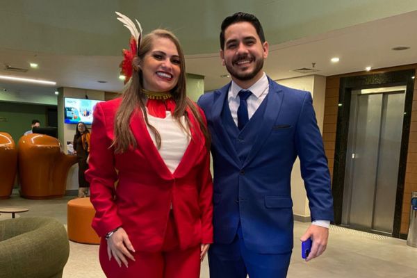 Uma mulher sorrindo, vestindo calça e blzaer vermelho, com camisa branca, abraçando um homem, vestindo terno azul, ambos sorrindo