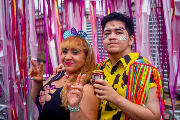 Uma mulher e um homem sorrindo, maquiados e vestidos com roupas alegres para o carnaval. O Homem segura uma lata de cerveja