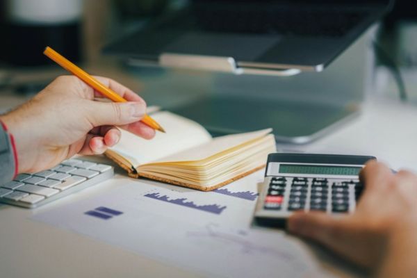 Aparece em destaque uma mão com um lápis, sobre um caderno e ao lado uma calculadora