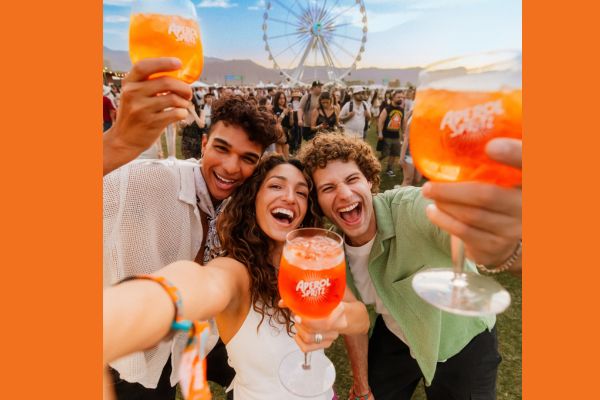 Dois homens e uma mulher ao centro, sorrindo e segurando uma taça com um drink Aperol Spritz, na cor laranja
