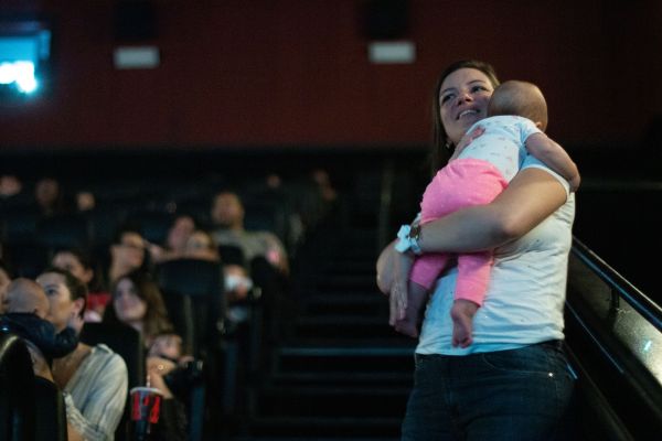Uma mãe, sorrindo, carregando seu filho no colo, em uma sala de cinema