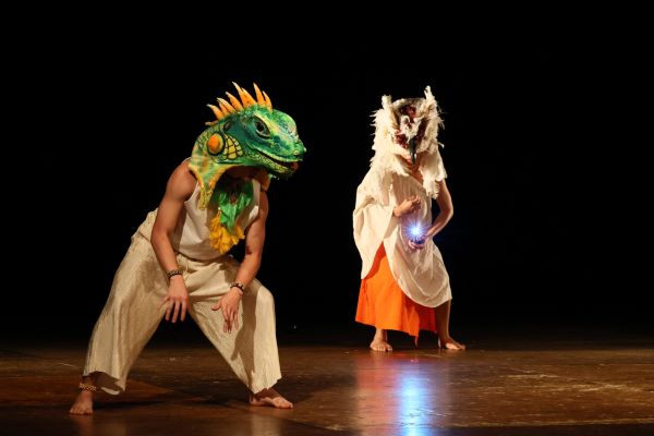 Personagens estão no palco em apresentação, um deles está com uma máscar em formato de lagarto