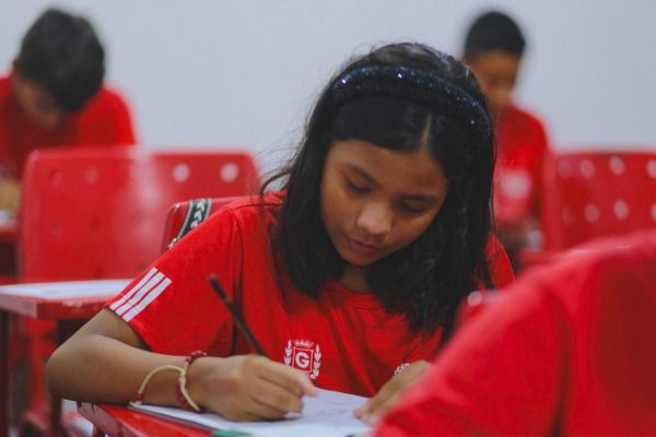 Uma menina estudando, com uma caneta na mão direita, vestindo uma camisa vermelha estilizada com o brasão do Boi Garantido
