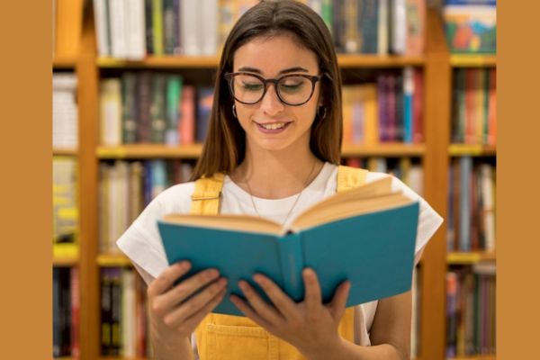 Uma jovem, de blusa branca e jardineira amarela, em uma biblioteca, sorrindo, fazendo a leitura de um livro que está em suas mãos