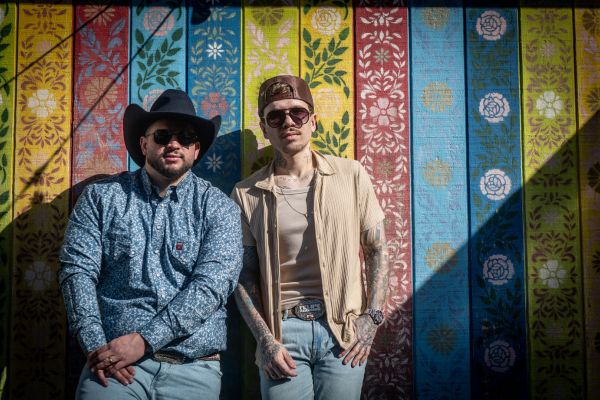 Felipe (de chapéu d ecouro) e Rodrigo (de chapéu marrom), ambos de pé, em uma parede multicolorida