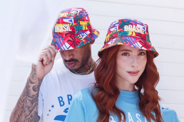 Um rapaz e uma moça, ambos estão usando um Buket colorido, com o nome Brasil e com estampas que valorizam a brasilidade e refletem a diversidade cultural do país
