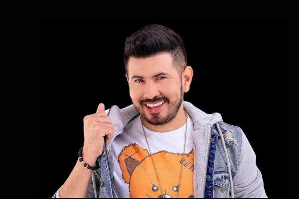 Cantor Japa, uma das atrações do evento, sorrindo, de blusa branca com a imagem de um urso, com uma jaqueta jeans claro