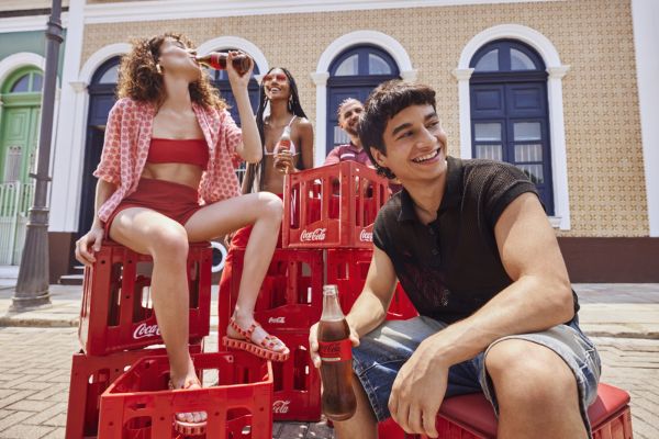 Quatro jovens, felizes e rindo, tomando coca-cola, uma mulher d evermelho sobre um engradado da marca e um homem de blusa preta, sobre outro