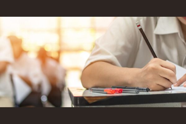 Em muma sala de aula, aparece parte do braço de uma estudante escrevendo e ainda uma visão de uma sala de aula, com alunos