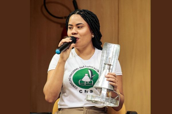 Letícia Santiago de Moraes autora do HQ vestindo camisa branca com destaque ao símbolo do Conselho Nacional das Populações Extrativistas (CNS)