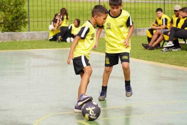 Duas crianças jogando futebol, vestindo camisa amarela e shorts preto